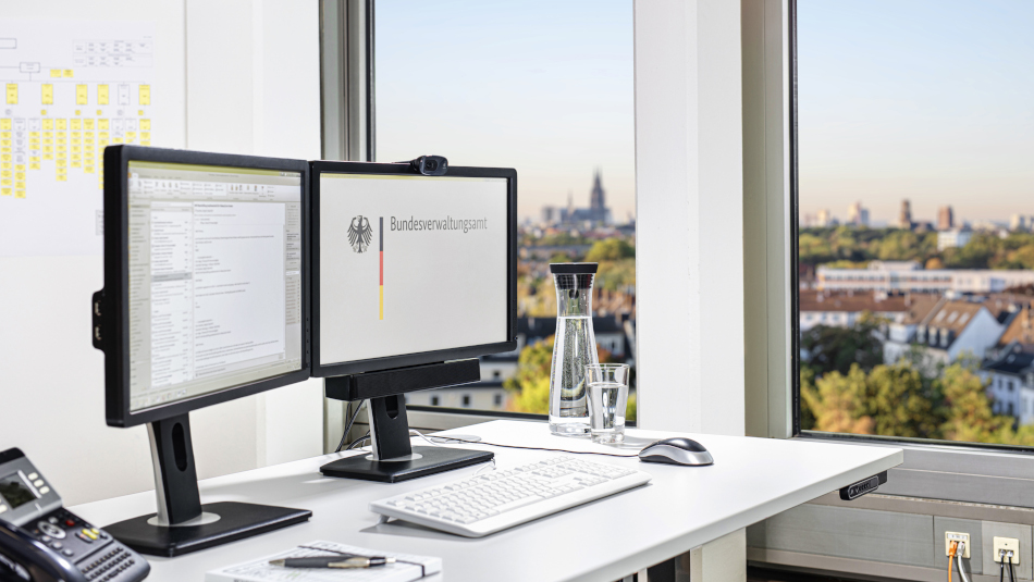 Schreibtisch mit zwei Bildschirmen, einer Tastatur und Computer-Maus, einem Telefon sowie ein Notizblock mit Stift. Vom Schreibtisch aus blickt man aus dem Fenster mit dem Kölner Dom im leicht verschwommenen Hintergrund.